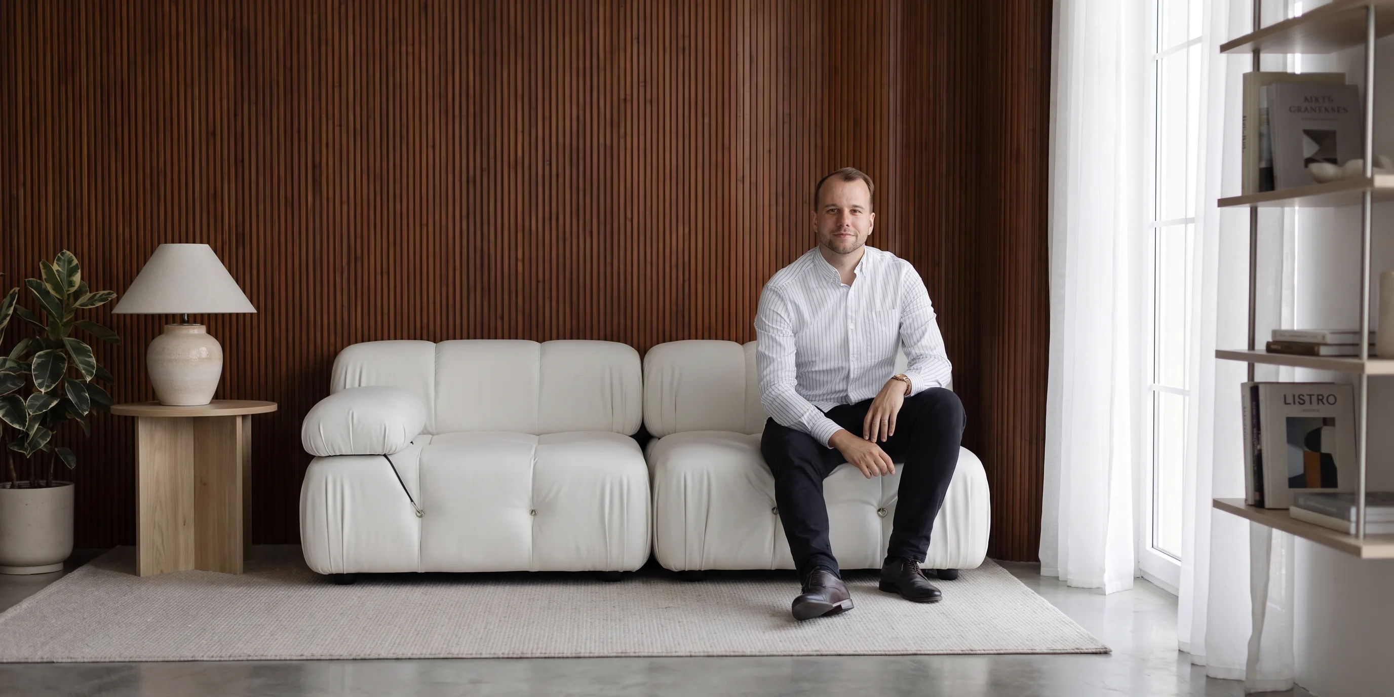 Chris P. sitting on a white sofa in a sunlit room with a slatted walnut wall behind him