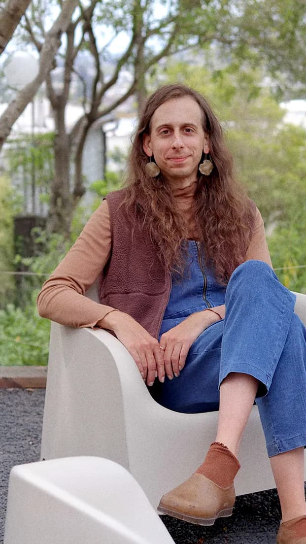 Lily Bodnar sitting in a white molded chair on a gravel patio, trees and a house in the background